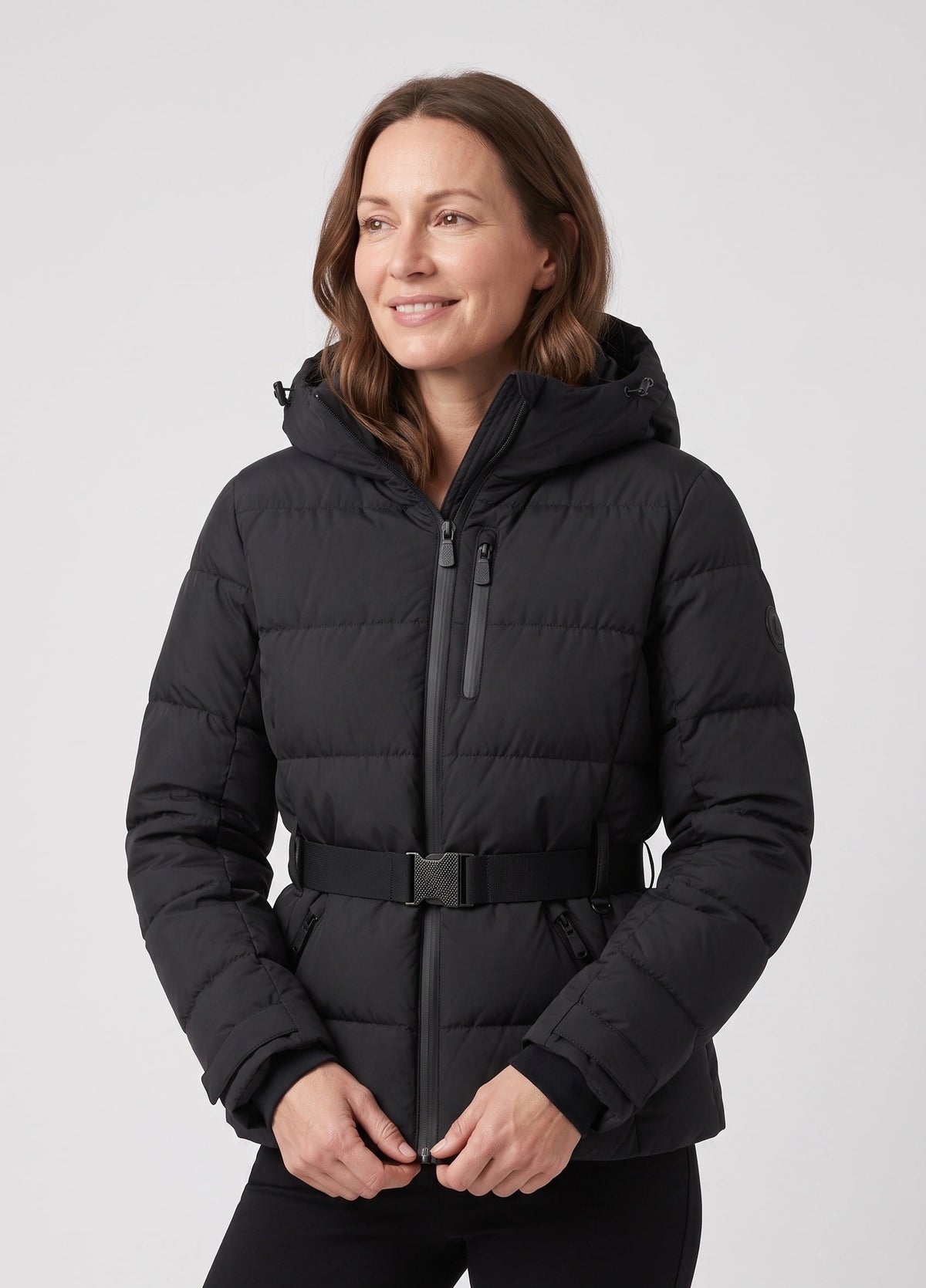 A woman wearing a black hooded puffer jacket with a front zipper, zip chest pocket, and a matching black belt, photographed against a light gray studio background.